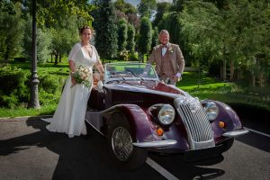 Photo de mariage : les mariés, élégamment vêtus, posent à côté d'une voiture de sport ancienne (vintage) de couleur bordeaux foncé, dans un parc verdoyant et ensoleillé.