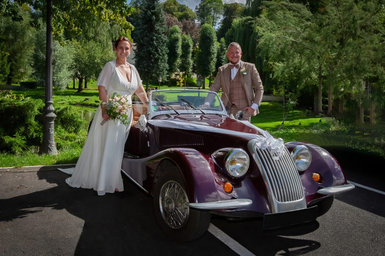 Photo de mariage : les mariés, élégamment vêtus, posent à côté d'une voiture de sport ancienne (vintage) de couleur bordeaux foncé, dans un parc verdoyant et ensoleillé.