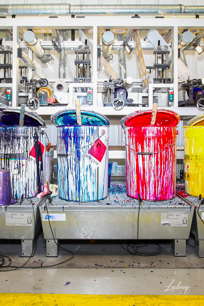 Quatre grands fûts industriels de peinture (bleu, rouge, jaune) débordants, avec des coulures de peinture intenses, posés sur un support métallique dans une usine. Des machines de production sont visibles en arrière-plan.
