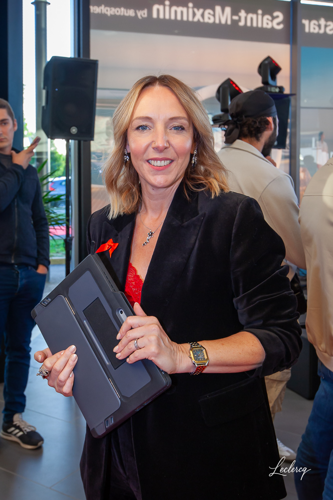 Portrait souriant d'une femme d'affaires blonde et élégante portant un blazer en velours noir et tenant une tablette, prise lors d'un événement corporate intérieur. Des invités sont visibles en arrière-plan flou.