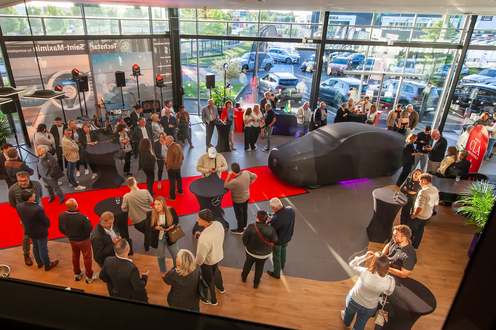 Vue intérieure de la concession Mercedes-Benz de Saint Maximin dans l'Oise lors de la soirée de lancement de la nouvelle CLA. La voiture est visible sous une bâche, entourée de convives élégants ou d'un groupe de personnes observant le véhicule. L'ambiance est festive et professionnelle.