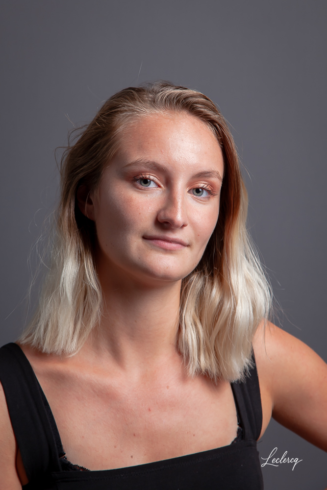 Portrait professionnel. Gros plan d'une jeune femme aux cheveux blonds mi-longs, posant de face sur un fond gris uni. Elle porte un débardeur noir et regarde l'objectif avec un sourire discret.