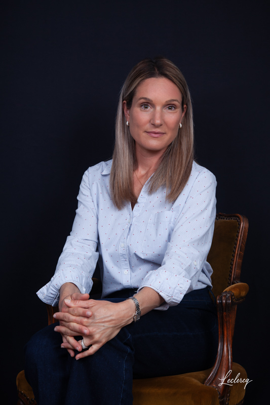 Portrait professionnel d'une femme blonde assise dans un fauteuil de style classique sur fond noir, portant une chemise claire à petits motifs et des mains croisées.