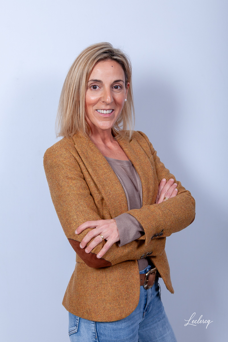 Portrait d'une femme souriante aux cheveux blonds mi-longs, posant avec les bras croisés devant un fond clair. Elle porte un blazer en tweed de couleur moutarde avec des coudières marron, un haut beige et un jean bleu clair.