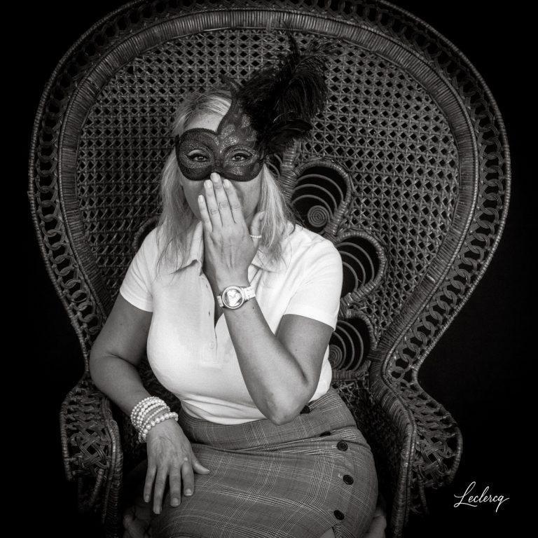 Portrait en noir et blanc d'une femme blonde assise dans un grand fauteuil en osier tressé. Elle porte un masque vénitien orné de plumes et place sa main devant sa bouche dans un geste de mystère. Elle porte également une montre, des perles au poignet et une jupe à carreaux.