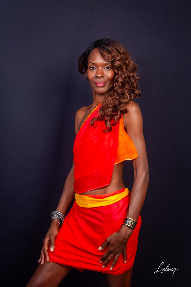 Portrait d'une femme souriante aux cheveux longs et bouclés, posant devant un fond noir. Elle porte une tenue asymétrique éclatante composée d'un haut et d'une jupe rouge avec des détails orange et jaune. Elle porte plusieurs bracelets larges aux poignets.