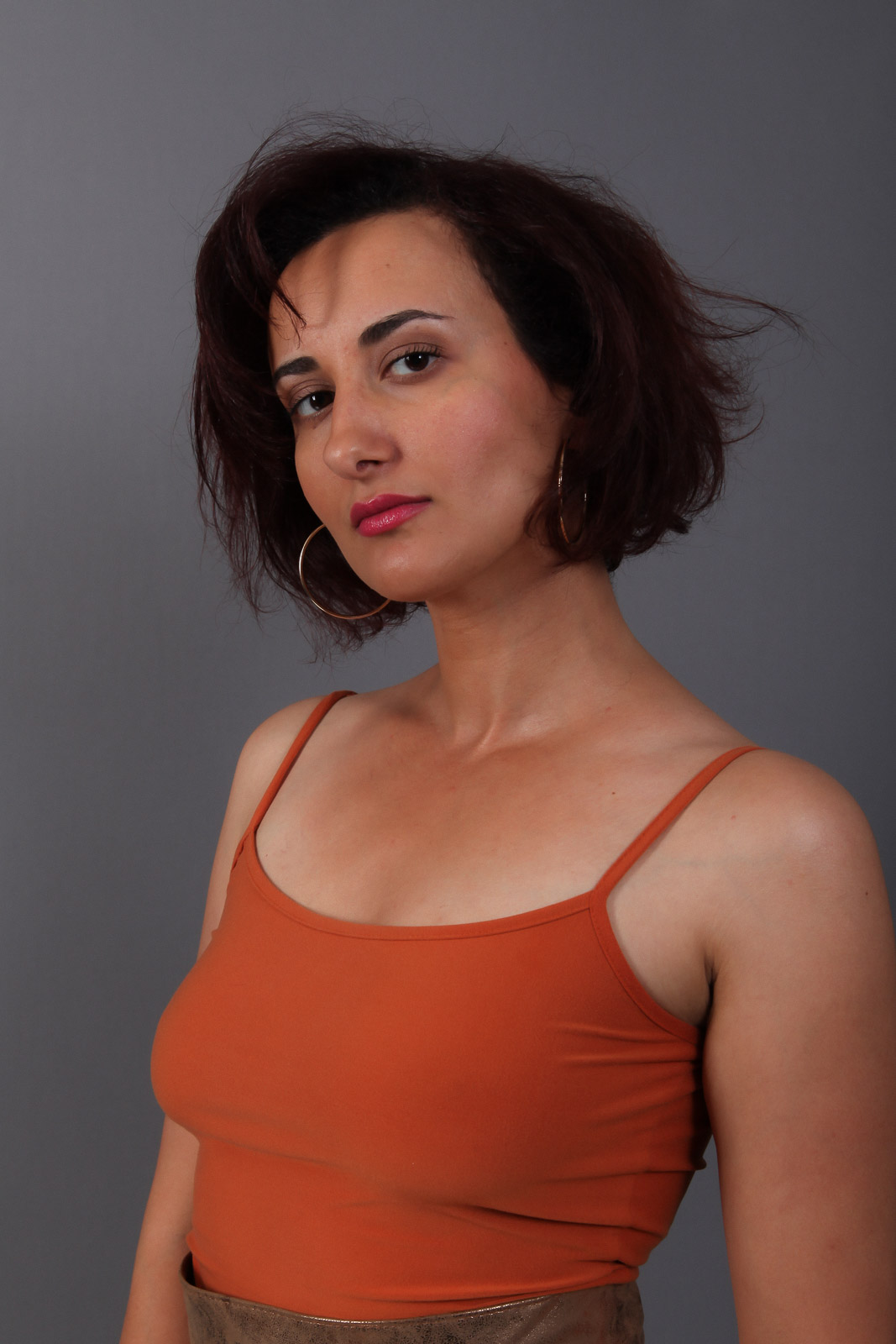 Portrait d'une femme aux cheveux courts bruns portant un caraco orange et des créoles dorées sur un fond gris.
