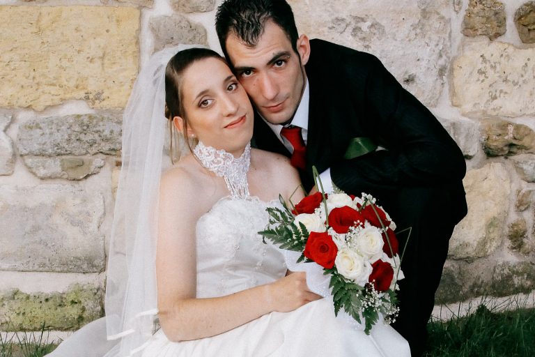 Portrait rapproché d'un couple de mariés assis devant un mur en pierre, la mariée tenant un bouquet de roses rouges et blanches.