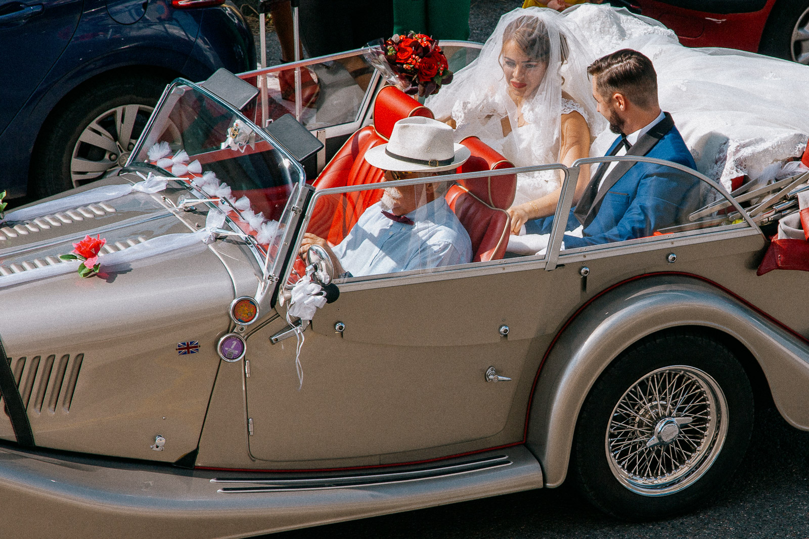 Mariés souriants à l'arrière d'une voiture de collection décapotable grise avec intérieur cuir rouge, conduite par un chauffeur avec un chapeau blanc.