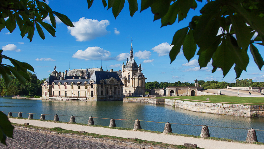 Stage photo Chantilly. Le Château entouré d'eau sous un ciel bleu, encadré par des feuilles d'arbres au premier plan.