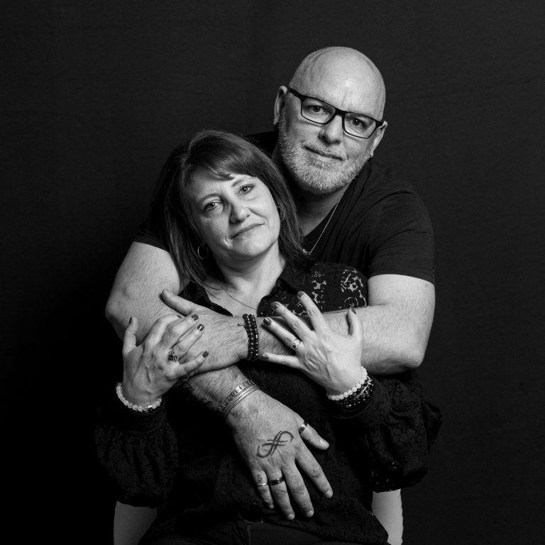 Portrait intimiste en noir et blanc d'un couple sur fond sombre. L'homme, portant des lunettes et barbu, enlace la femme par derrière. Leurs mains entrelacées mettent en avant des bijoux et un tatouage de l'infini.
