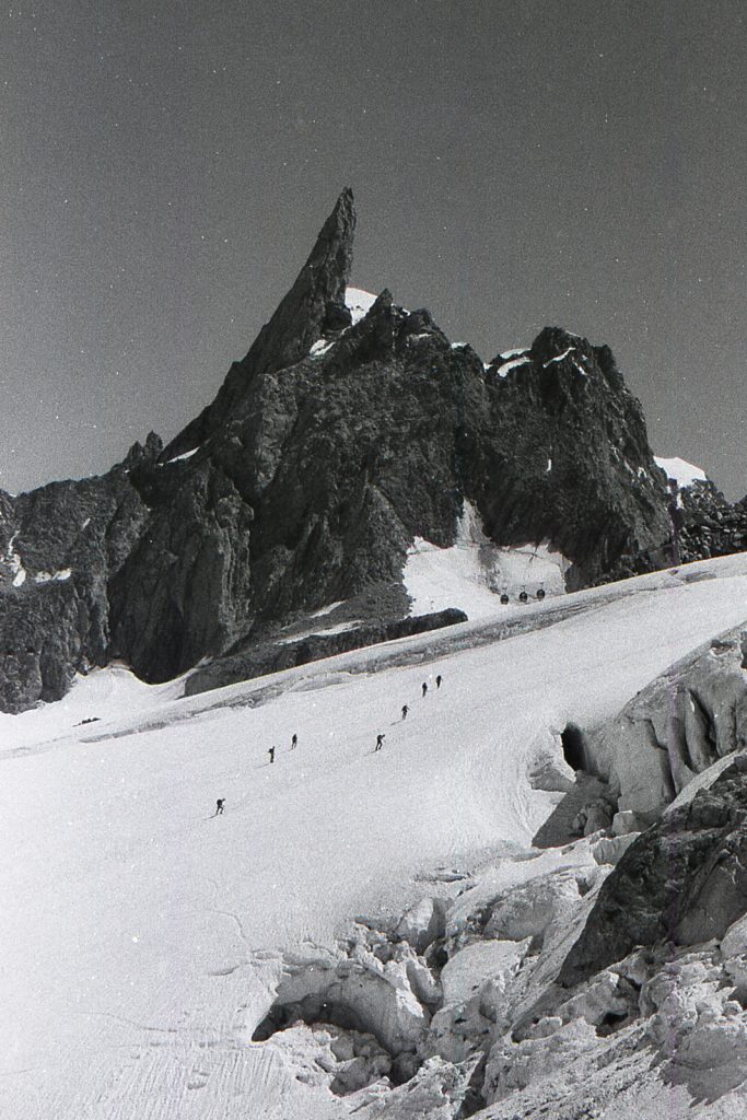 Photo argentique en noir et blanc. La Dent du Géant vue depuis le Glacier du Géant. Sept alpinistes remonte vers la pointe Helbronner. On peut apercevoir les télécabines.