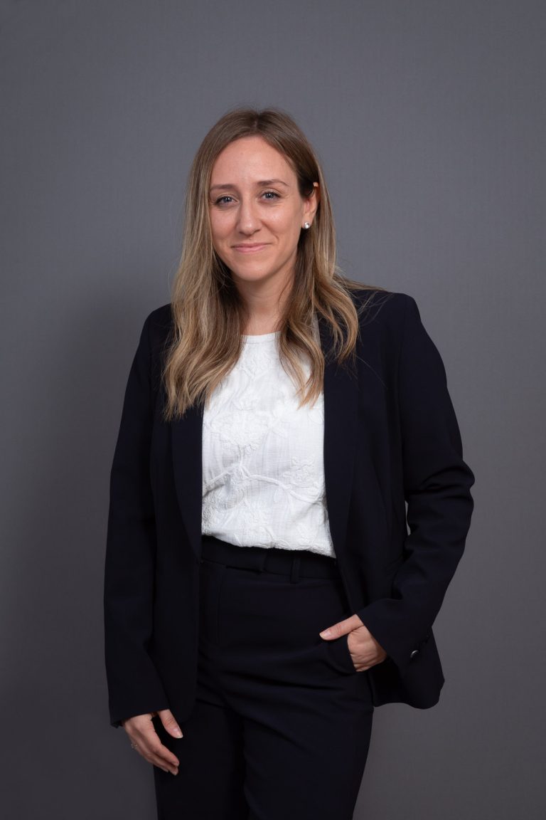 Portrait en plan moyen d'une femme souriante en tailleur sombre et haut blanc, une main dans la poche, posant devant un fond gris neutre.