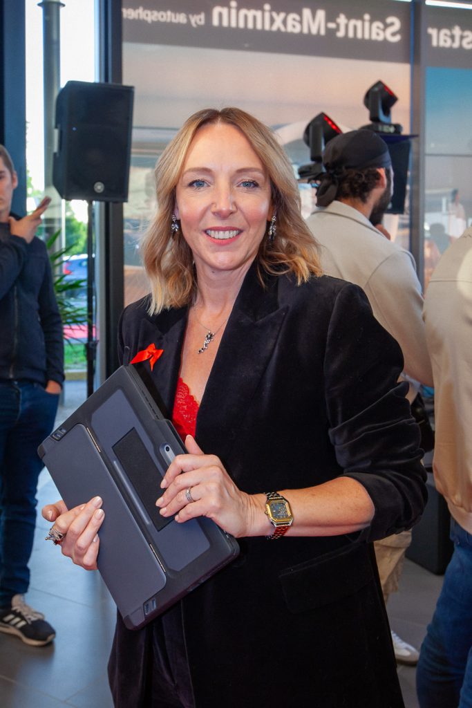 Portrait en plan moyen d'une femme blonde souriante tenant une tablette tactile, vêtue d'une veste noire lors d'un événement corporate en intérieur.