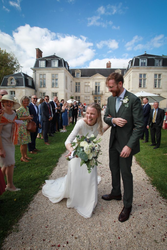 Mariée souriante en robe blanche avec bouquet et marié en costume vert marchant sur une allée en gravier, entourés d'invités devant un château en pierre blanche.